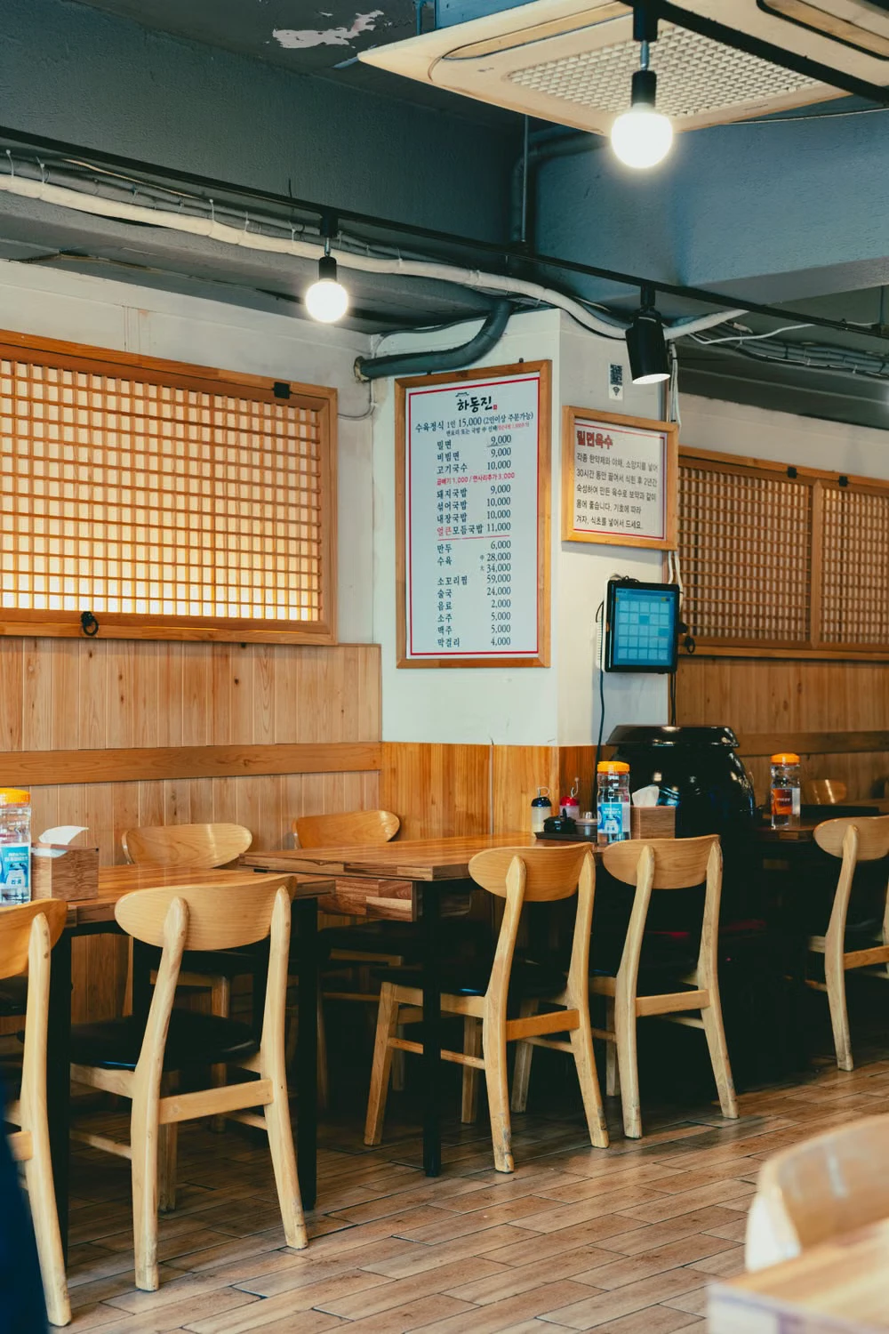 A casual Korean restaurant interior with open wooden tables, a wall menu, and an ordering screen