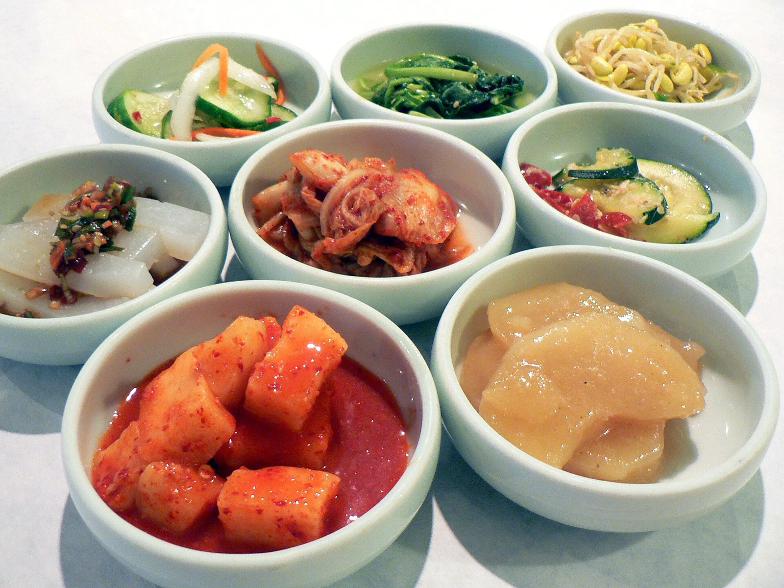 Eight bowls of Korean banchan including kimchi, radish, greens, cucumber, and bean sprouts