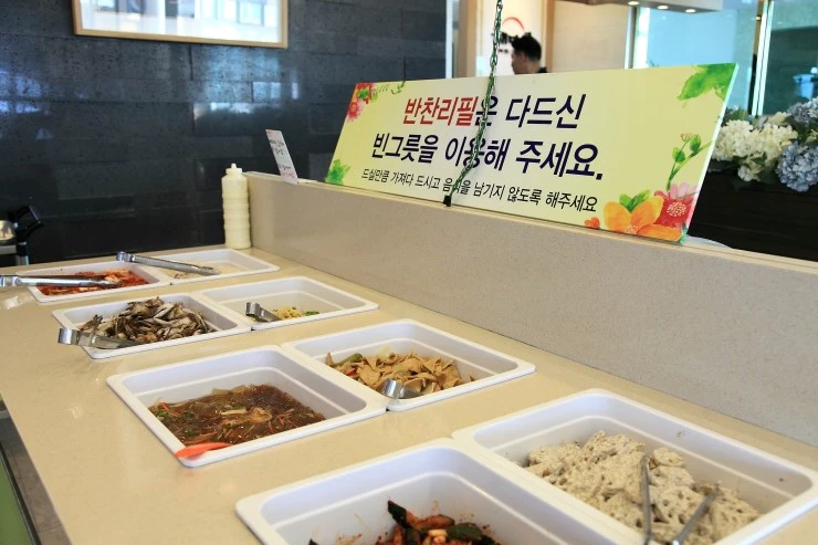 A Korean restaurant self-service banchan bar with separate trays, serving tongs, and a sign asking diners to take only what they will eat