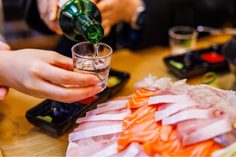 Clear soju being poured from a green bottle beside a sashimi meal