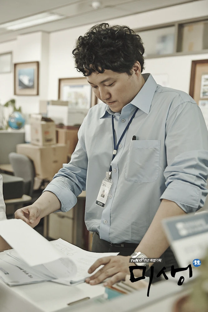 An office employee reviewing documents at a shared workstation in the tvN drama Misaeng