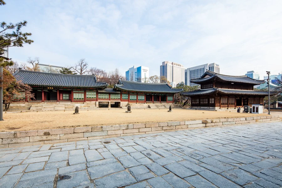 Traditional palace buildings and a modern skyline across a stone court at Deoksugung