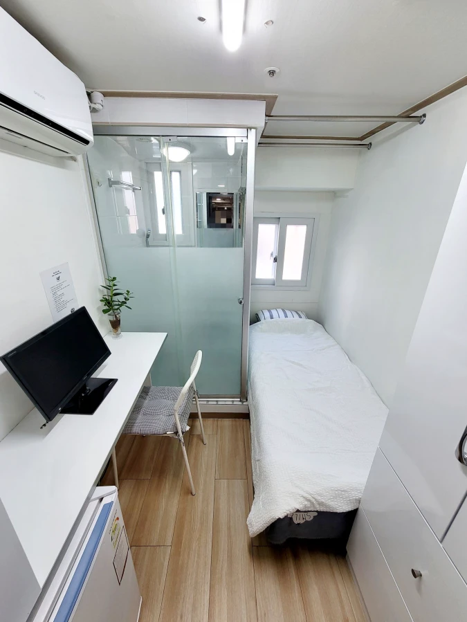 Compact renovated goshiwon room with a single bed, desk, refrigerator, air conditioner, and private shower