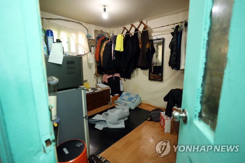 Older lived-in Seoul goshiwon room with floor bedding, refrigerator, clothes rack, and very limited open space