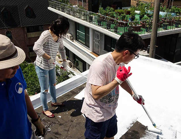 Volunteers applying white heat-reflective coating to a rooftop beside a Korean rooftop room