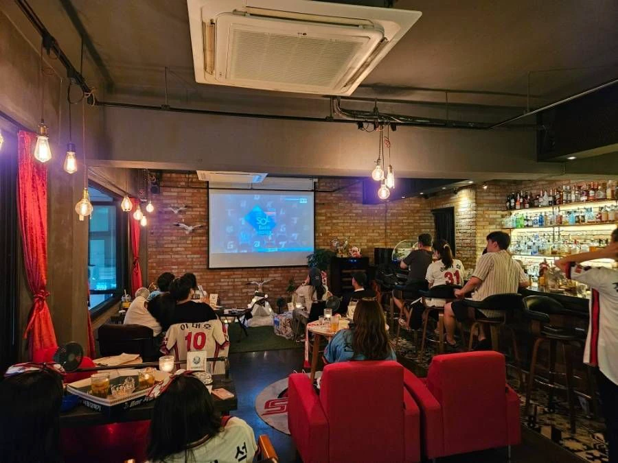 Baseball fans in team jerseys watching a game on a large screen inside a Korean baseball pocha