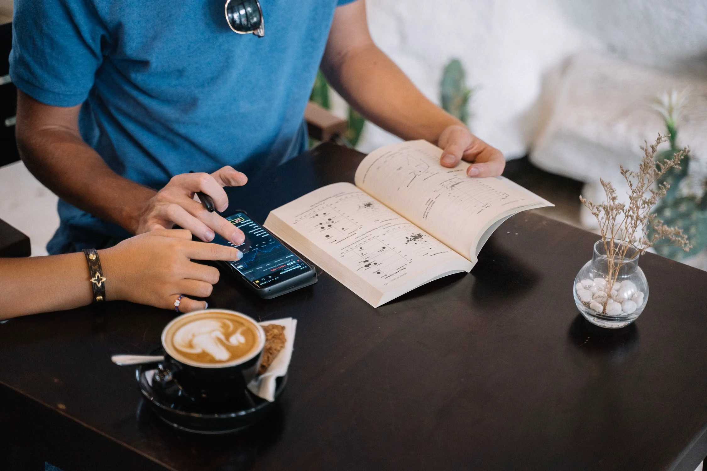 Dua orang membandingkan ponsel dan buku terbuka sambil menikmati kopi di kafe