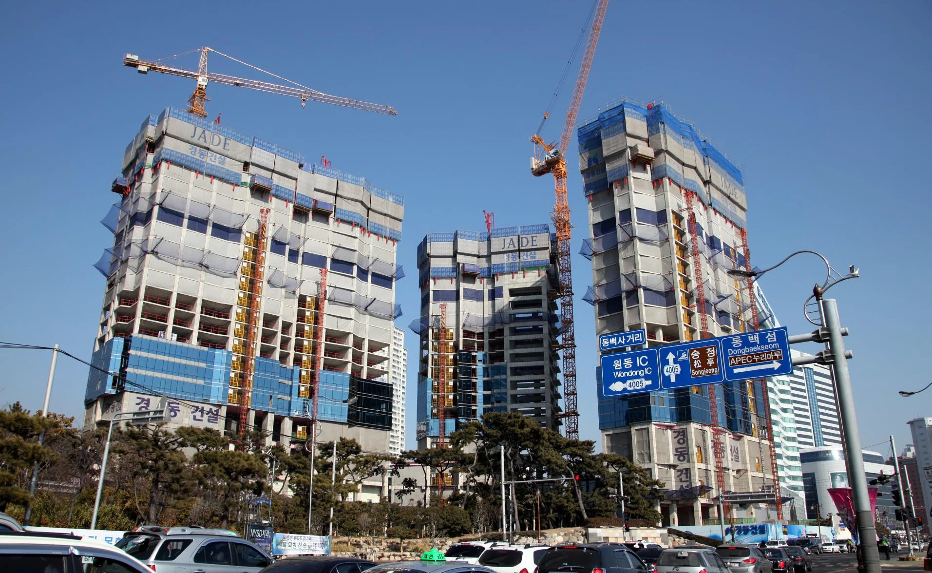 Three Korean high-rise residential towers rising behind cranes and protective construction screens