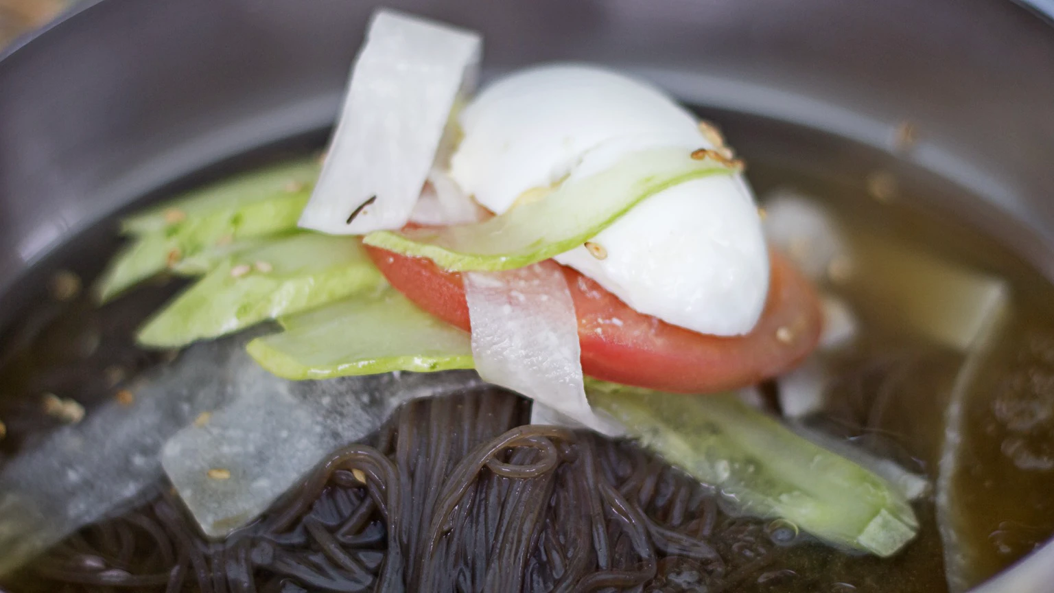 Close view of chilled naengmyeon noodles with cucumber, egg, radish, and tomato in broth