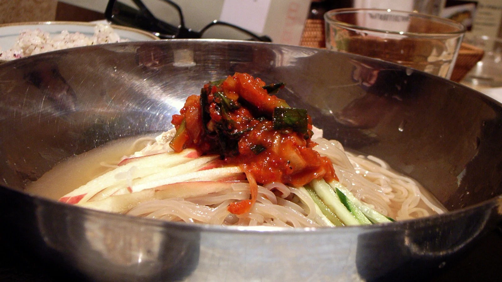 Metal bowl of cold naengmyeon with translucent noodles, shredded fruit, cucumber, and spicy garnish