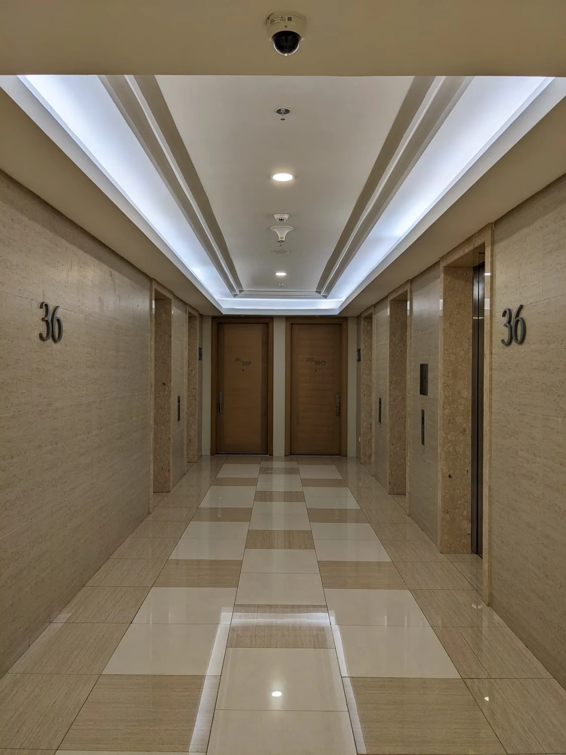 Bright hotel corridor with elevators, room doors, ceiling lights, and a visible security camera