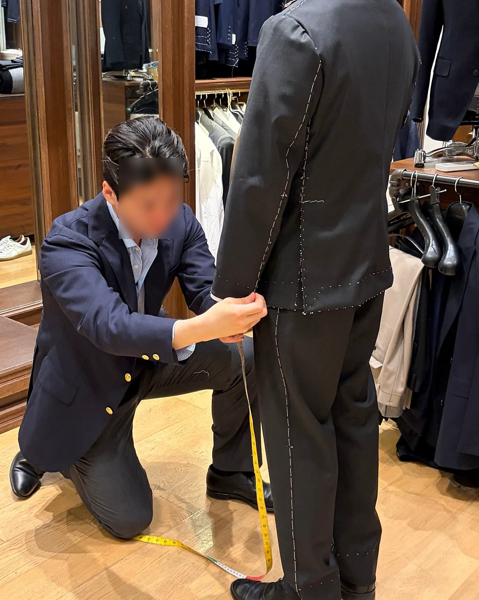 Tailor measuring the jacket hem during a basted fitting while the customer stands in front of a mirror