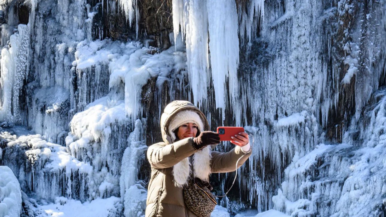 Traveler in a hooded winter coat taking a photo in front of the frozen Hongjecheon Artificial Waterfall in Seoul