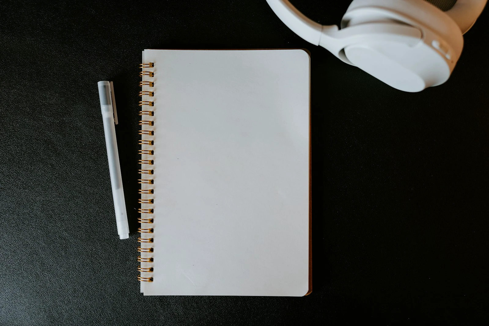 Buku catatan kosong, pulpen, dan headphone di atas meja berwarna gelap