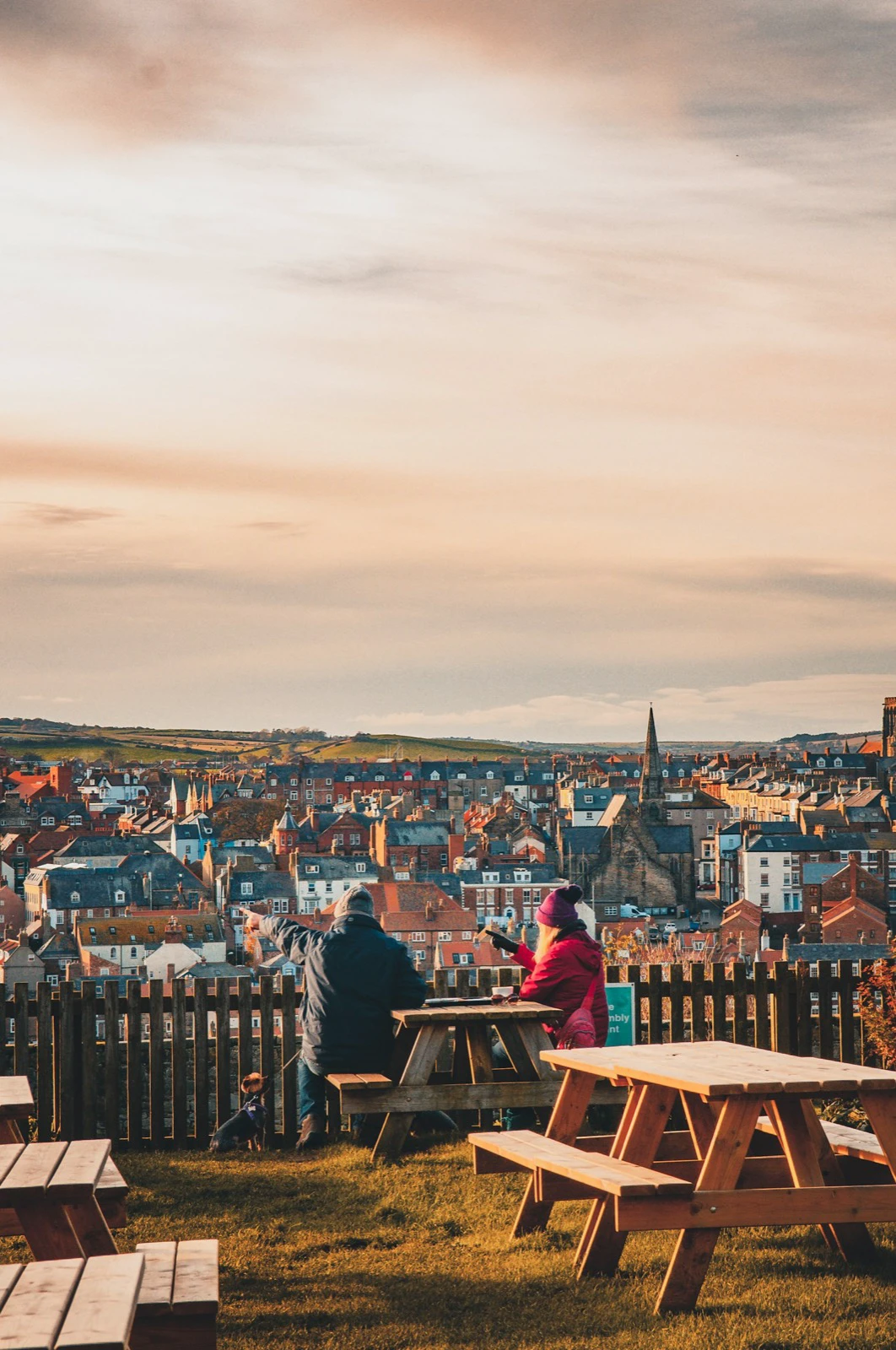 Two people in warm clothing seated at an outdoor picnic table overlooking a town