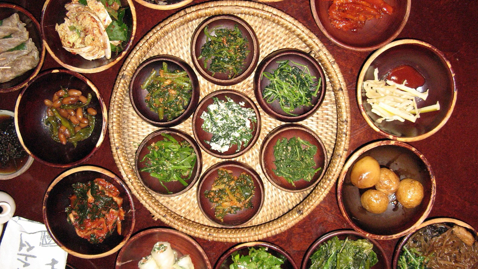 Overhead spread of Korean temple dishes arranged in brown bowls around a woven tray
