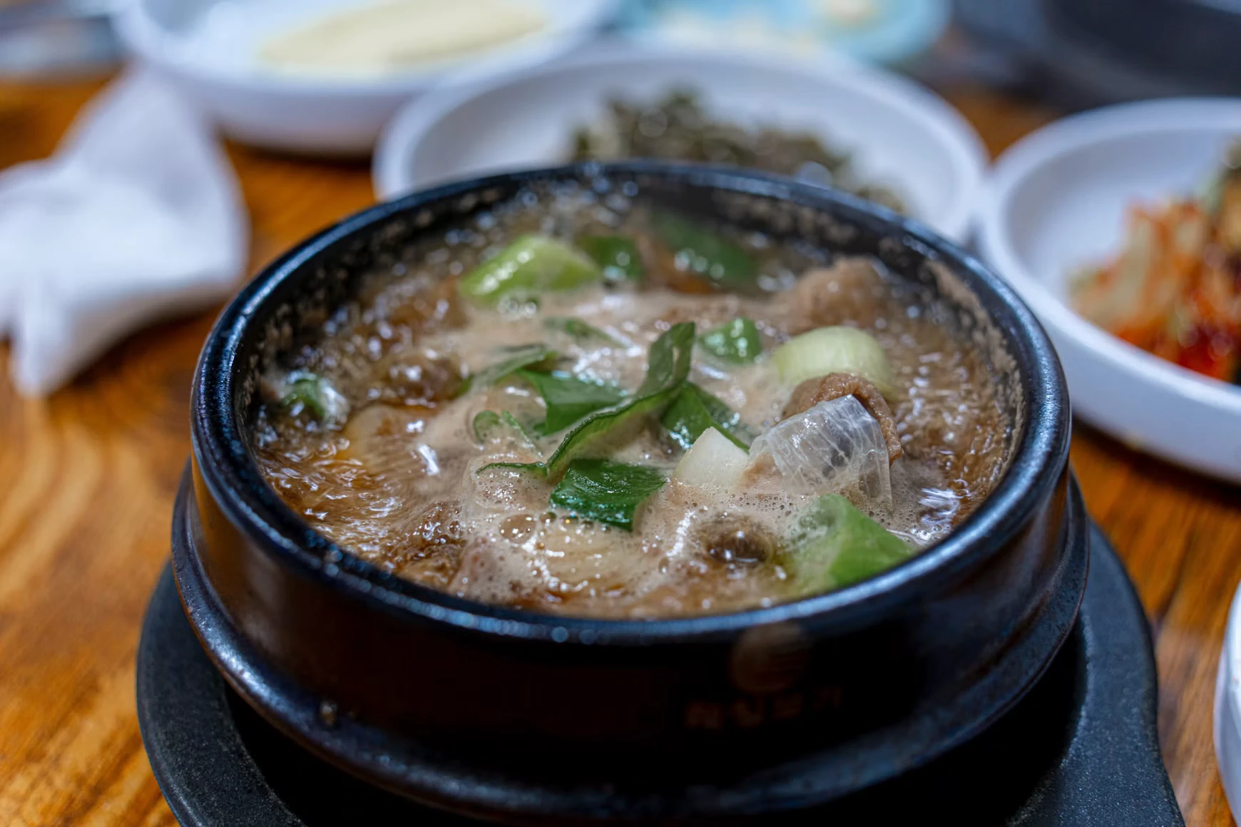 A bubbling Korean soup in an earthenware bowl with rice and side dishes behind it