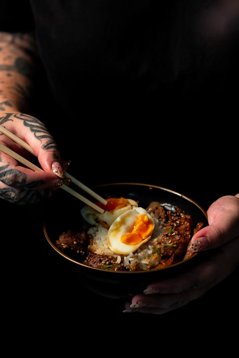 Hands holding a rice bowl topped with beef and a soft-boiled egg