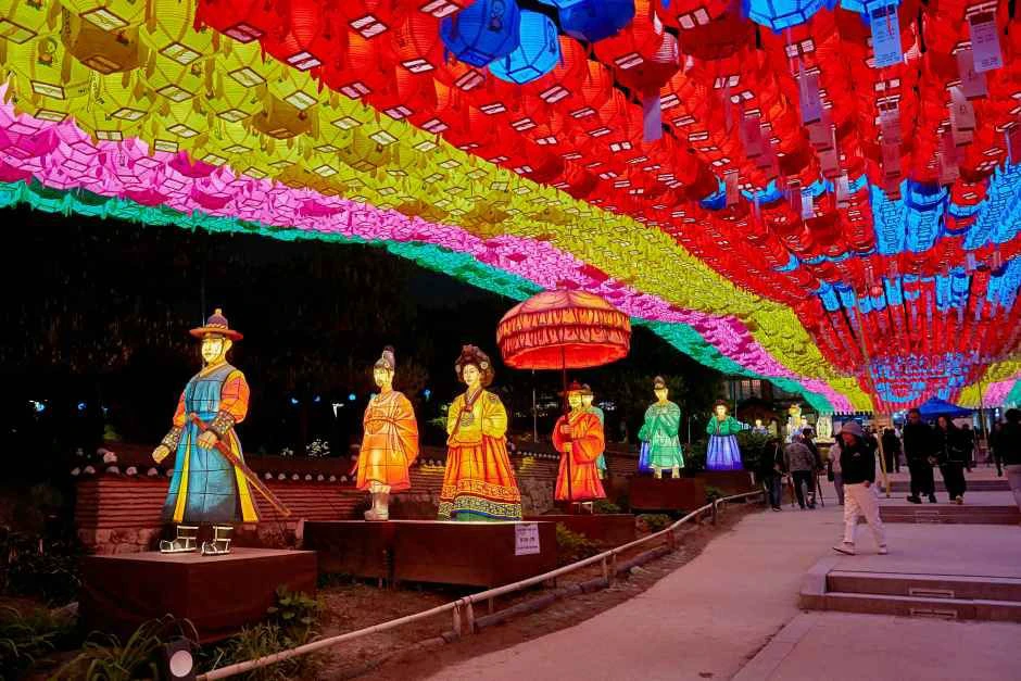A temple walkway covered by rows of colorful lotus lanterns beside illuminated traditional figures at night