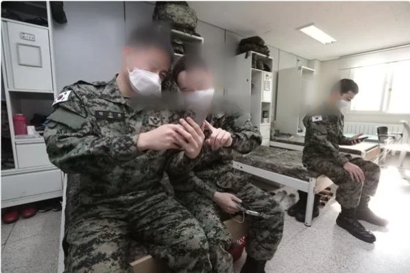 Two South Korean soldiers share a smartphone in their barracks while a third sits nearby.