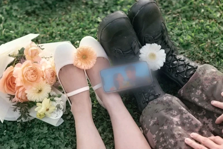 White shoes with flowers beside black military boots on grass; a phone screen is privacy-blurred.