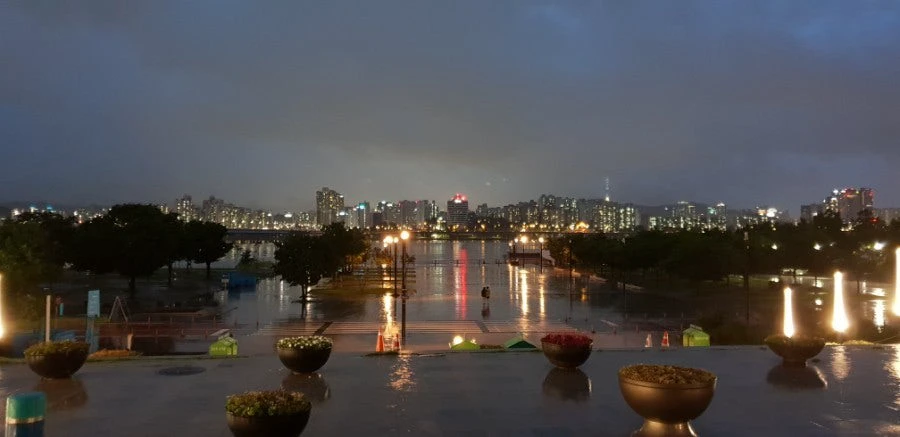 City lamps and skyline lights reflected across a rain-covered plaza after dark